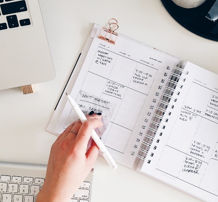 Photo of a woman's hand holding a pen, placing a sticky note on a paper planner with a gold binder clip at the top